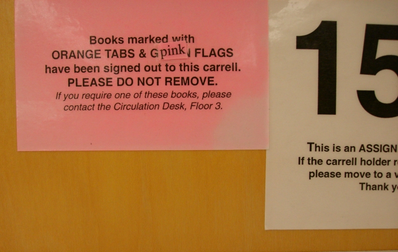 Sign explaining that books with orange tabs or pink flags have been checked out to the carrel and should not be removed. The word 'pink' is on a separate piece of paper and taped over another word that must have started with 'G'.