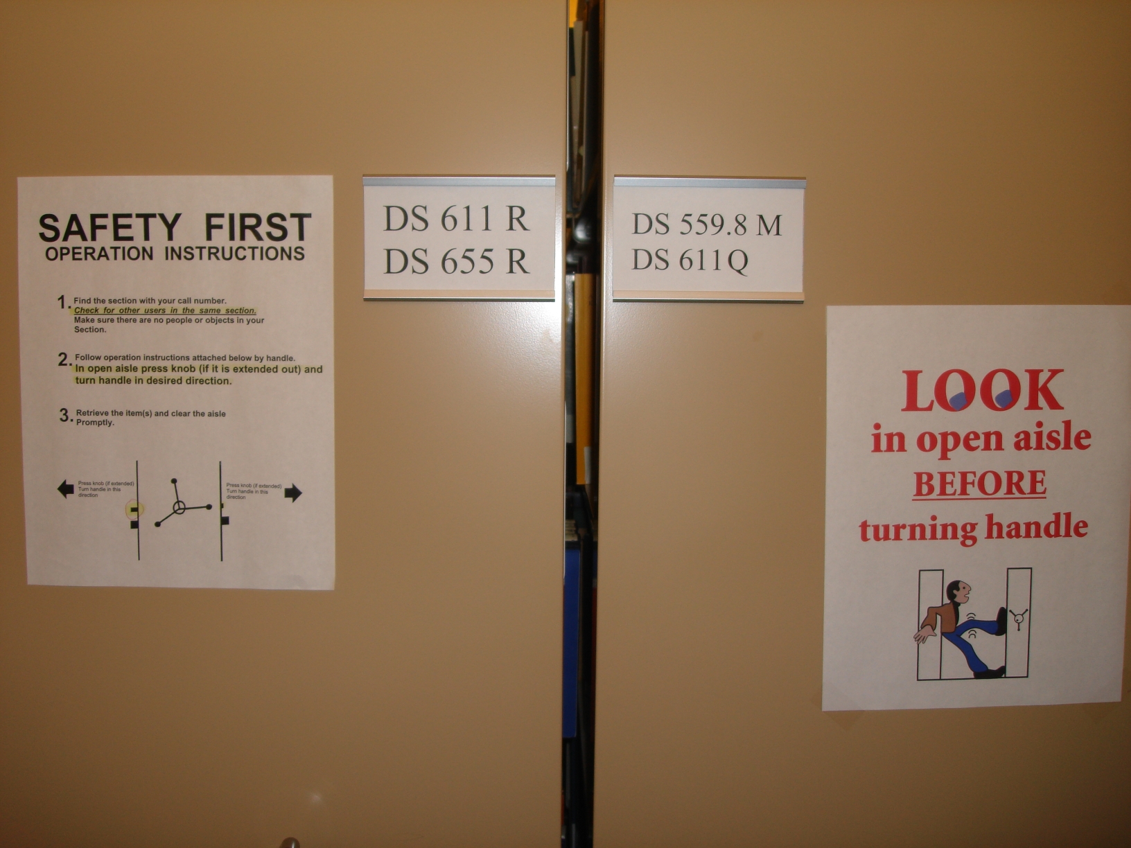 Side view of movabale shelves showing signage warning people to operate the shelves safely. One sign shows a person trying not to be crushed between two shelves. The wording says: "LOOK in open aisle BEFORE turning handle"