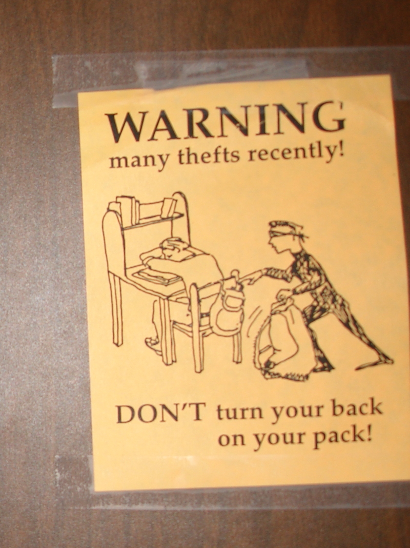 Sign warning you not to turn your back on your backpack because someone might steal it. Picture shows a cartoonish robber wearing black stealing from a student sleeping at a desk.