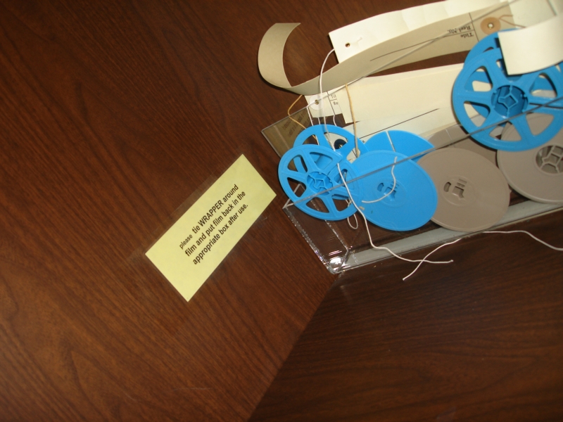 Picture of a desk with a sign on it asking people to rewrap microfilm before putting it back. There are some empty reels in the background.